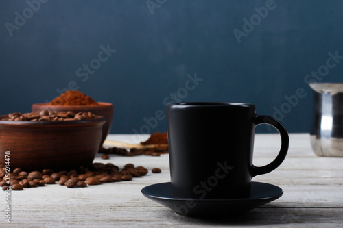 Grey cup of coffee, roasted Arabica beans in clay bowls, coffee powder, metal Turkish pot on while table against dark blue wall. Side view, copy space. Coffee shop, morning, baristas workplace concept