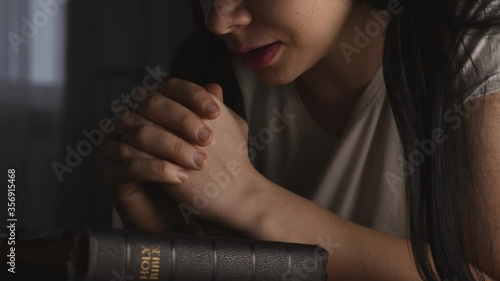 A woman prays with her hands crossed over a Bible for God's blessing in a dark room.