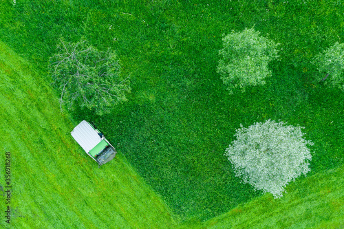 Lawn mower machine rides on grass, mowing tall grass in a city park among trees, aerial top view.