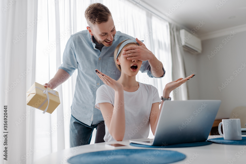 Fototapeta premium handsome man covering eyes of surprised girlfriend while holding present