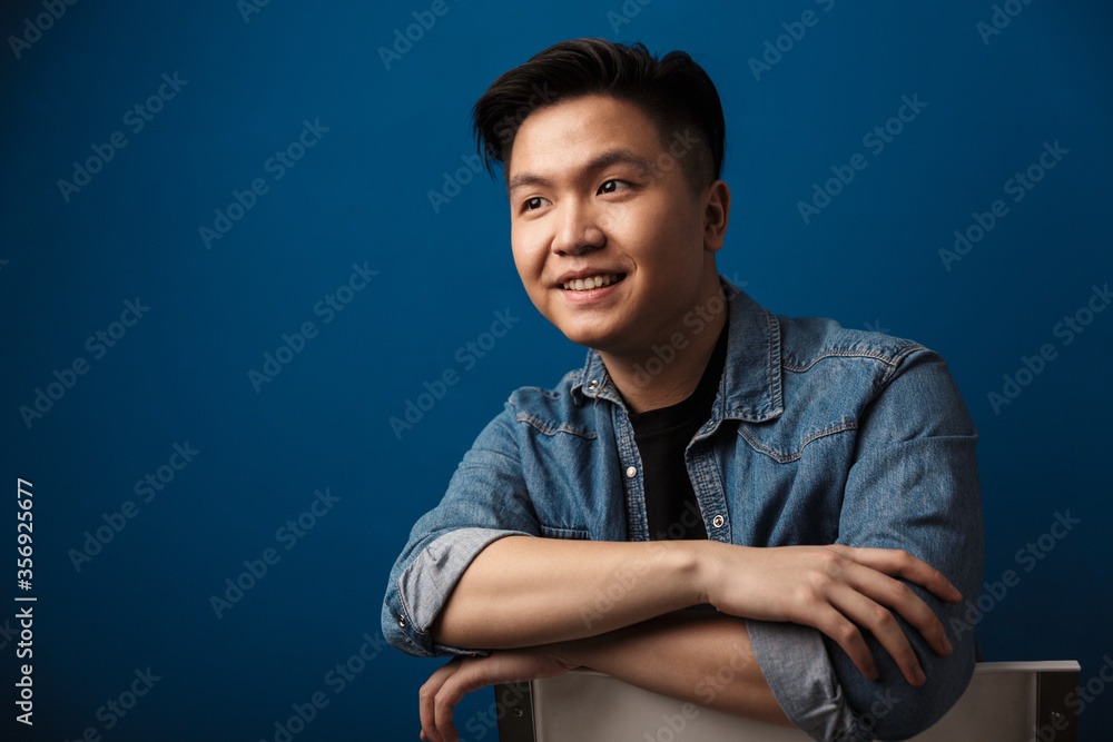 Image of excited handsome asian man smiling while sitting on chair