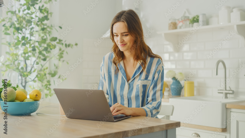 Young Beautiful Woman Using Laptop Computer While Wearing Blue Pyjamas. Brunette Female Sitting in a Modern Kitchen Room. Working from Home During Quarantine. Young Beautiful Woman Using Laptop Computer While Wearing Blue Pyjamas. Brunette Female Sitting in a Modern Kitchen Room. Working from Home During Quarantine.
