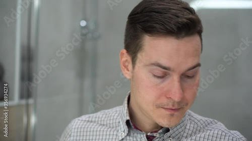 Young handsome Caucasian man in plaid shirt looks in mirror rubbing, massaging itchy brown eyes, puts on black glasses on gray background. Feeling tired and stressed. Problem with eyesight, vision.