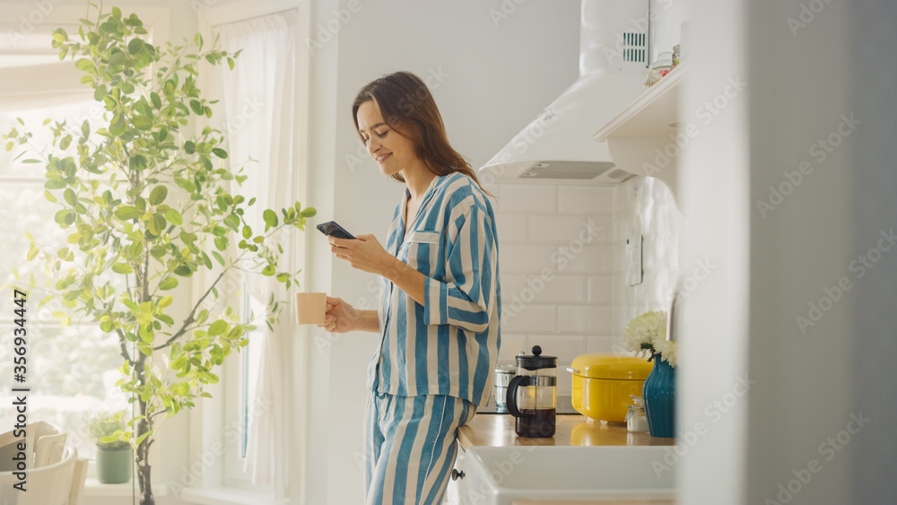 Beautiful Female is Using a Smartphone in a Kitchen While Drinking a Cup of Freshly Brewed Coffee. Girl in Pyjamas with Healthy Lifestyle Relaxes at Home in the Morning. Beautiful Female is Using a Smartphone in a Kitchen While Drinking a Cup of Freshly Brewed Coffee. Girl in Pyjamas with Healthy Lifestyle Relaxes at Home in the Morning.