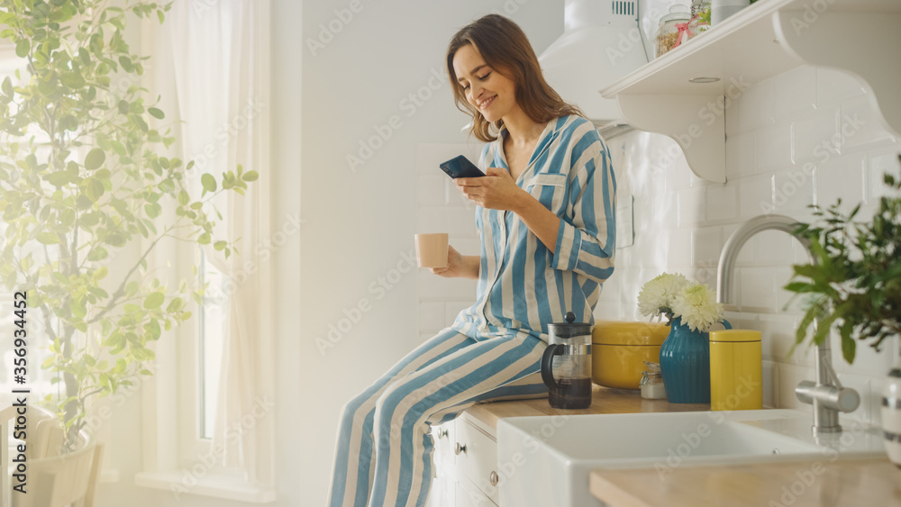 Beautiful Female is Sitting on a Cupboard and Using a Smartphone in a Kitchen While Drinking a Cup of Freshly Brewed Coffee. Girl in Pyjamas with Healthy Lifestyle Relaxes at Home in the Morning.