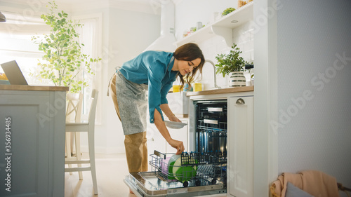 Beautiful Female is Loading Dirty Plates into a Dishwasher Machine in a Bright Sunny Kitchen. Girl in Wearing an Apron. Young Housewife Uses Modern Appliance to Keep the Home Clean. photo