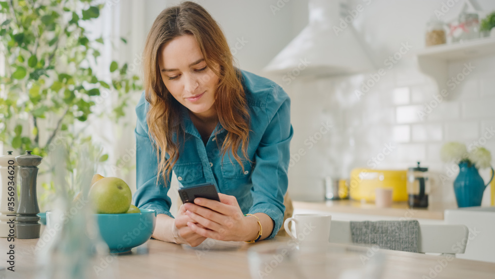 Beautiful Female is Using a Smartphone in a Kitchen. Pretty Young Woman Poses for the Camera and Gently Smiles. Attractive Girl with Healthy Creative Lifestyle Relaxes at Home in the Morning. Beautiful Female is Using a Smartphone in a Kitchen. Pretty Young Woman Poses for the Camera and Gently Smiles. Attractive Girl with Healthy Creative Lifestyle Relaxes at Home in the Morning.