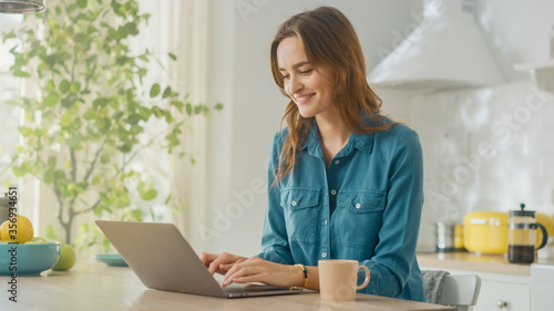Young Attractive Woman Using Laptop Computer and Drinking Morning Cup of Coffee While Wearing a Jeans Shirt. Brunette Female Sitting in a Modern Cozy Kitchen. Freelancer Working from Home. © Gorodenkoff