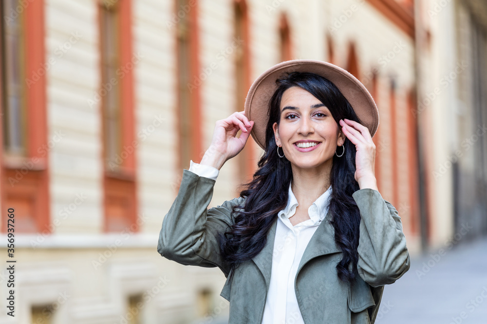 Fototapeta premium Outdoor portrait of a young beautiful fashionable happy lady posing on a street of the old city. Model wearing a hat. Girl looking up. Female fashion. City lifestyle. Copy space for text