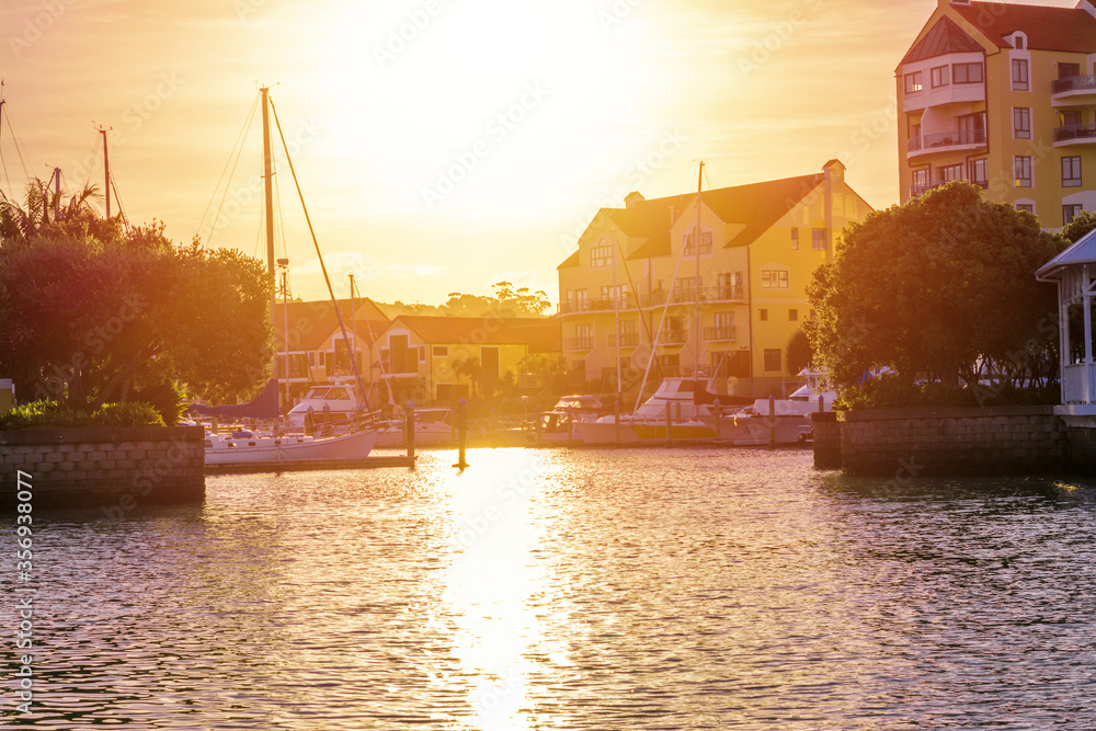 Naklejka premium Golden sun setting behind picturesque house by Gulf Harbour town basin. Sailboats moored nearby, Auckland, New Zealand