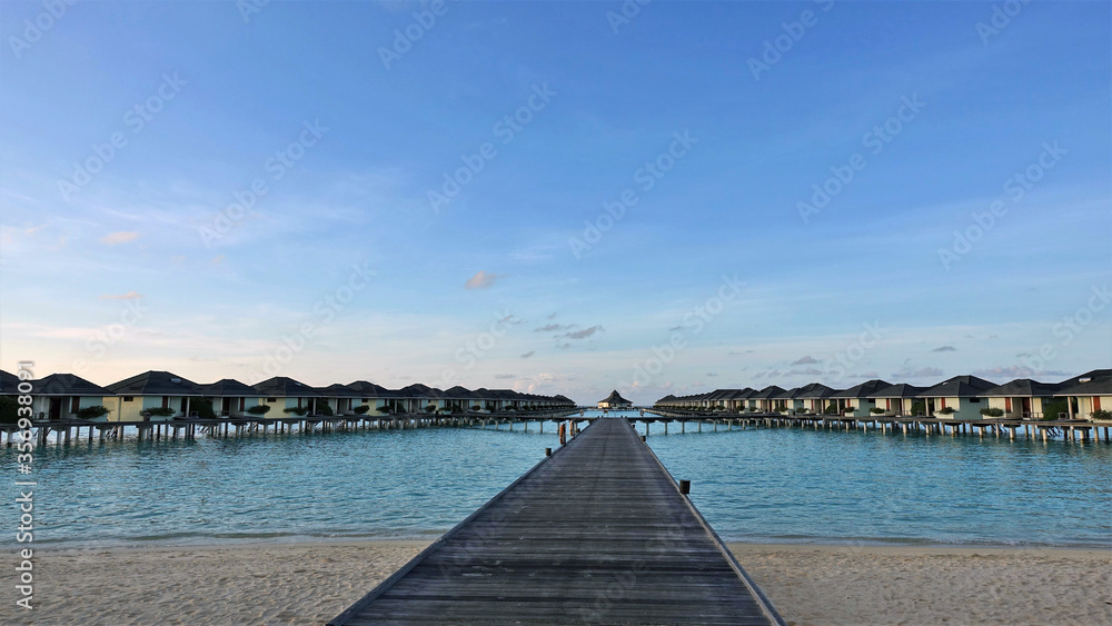 A clear sunny day in the Maldives. A wooden platform goes from the sandy shore to the villas on the water. Calm aquamarine ocean, bright blue sky. Silence and happiness.