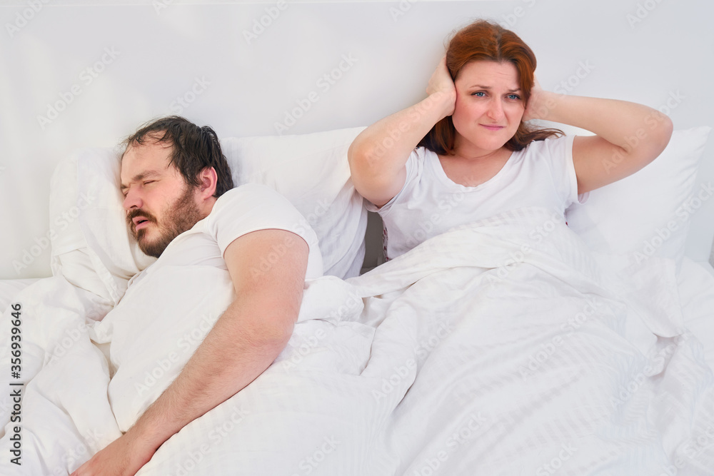 A woman puts her hands over her ears because of the noise and snoring of a man. Husband and wife spend time at home under the coronavirus quarantine. A woman puts her hands over her ears because of the noise and snoring of a man. Husband and wife spend time at home under the coronavirus quarantine.
