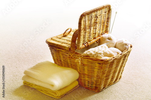 A pile of folded knitware by a wicker basket full with yarn roll, knitting accessories and knitwork in progress. Selective focus. Light background photo