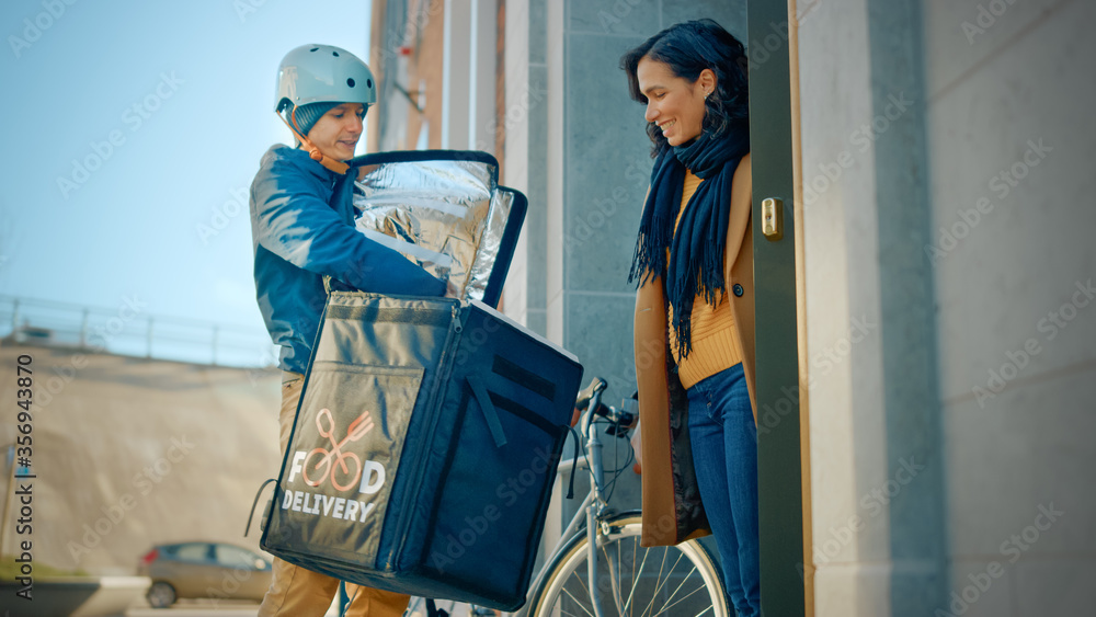 Happy Food Delivery Man Wearing Thermal Backpack on a Bike Delivers Restaurant Order to a Female Customer. Courier Delivers Takeaway Lunch to a Girl in Modern City District Office Building. Low Angle