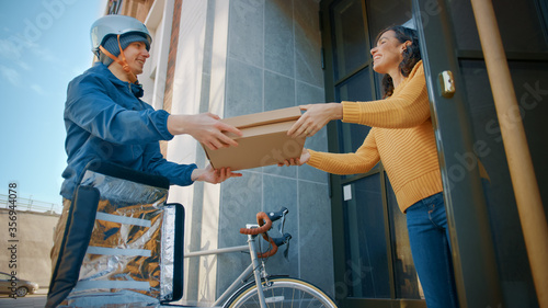 Happy Food Delivery Man Wearing Thermal Backpack on a Bike Delivers Pizza Order to a Beautiful Female Customer. Courier Delivers Takeaway Fast Food to a Smiling Girl in Office Building. Low Angle