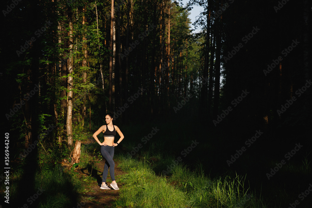 A beautiful woman in leggings and a top stands in the sunlight in the morning in a summer forest A beautiful woman in leggings and a top stands in the sunlight in the morning in a summer forest