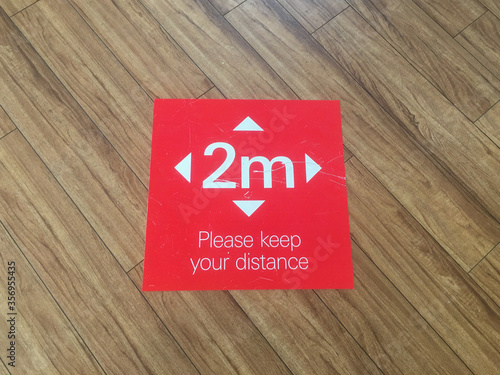 A floor sign with white lettering on red background says 'Please keep your distance'.Arrows point in four directions.Image