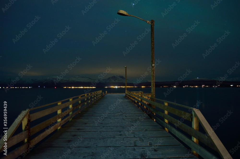 long snowy wooden pier with faint aurora borealis over mountain and fjord surface