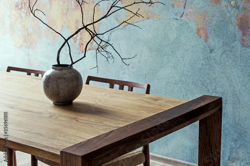 Close up of design wooden table with, stylish chairs, flowers in vase and elegant accessoreis. Details of home decor. Indusrtial concept of dining room. Open space. Template. Old wall.