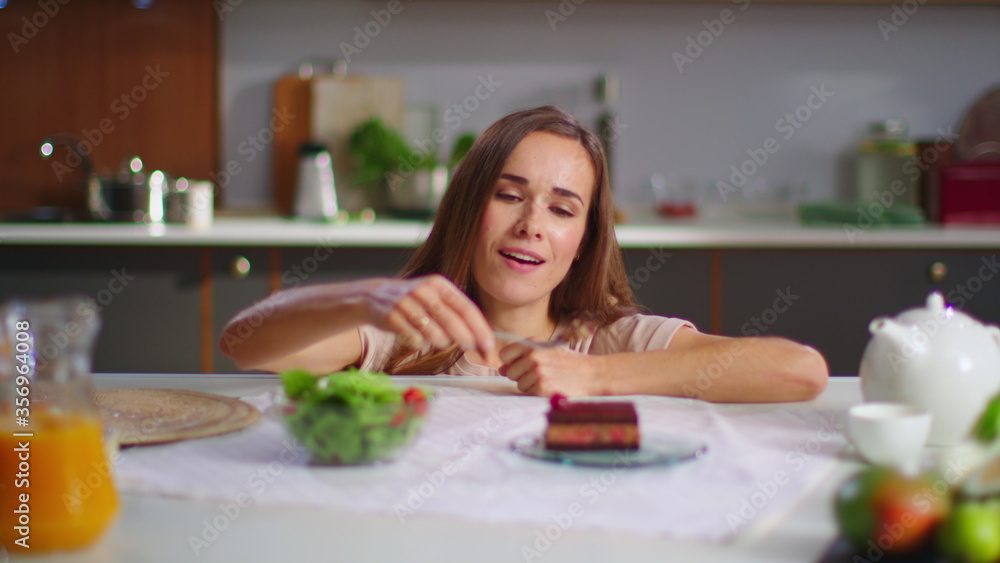 Happy woman eating cake instead salad on kitchen. Lady enjoying cake at table