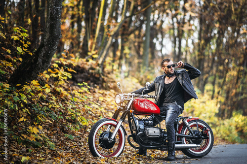 Bearded brutal man in sunglasses and leather jacket sitting on a motorcycle on the road in the forest