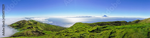 Panoramic view of green landscape above the clouds with Pico mountain in the distance, grass and ocean at the island of Sao Jorge, Azores, Portugal
