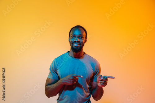 Pointing at side. African-american man's modern portrait on gradient orange studio background in neon light. Beautiful afro model. Concept of human emotions, facial expression, sales, ad. Copyspace.