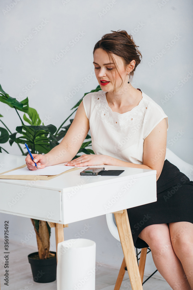 An elegant girl in a white shirt and black skirt sits at a white table. She thought. The girl sits in an office decorated in white and beige colors. An elegant girl in a white shirt and black skirt sits at a white table. She thought. The girl sits in an office decorated in white and beige colors.