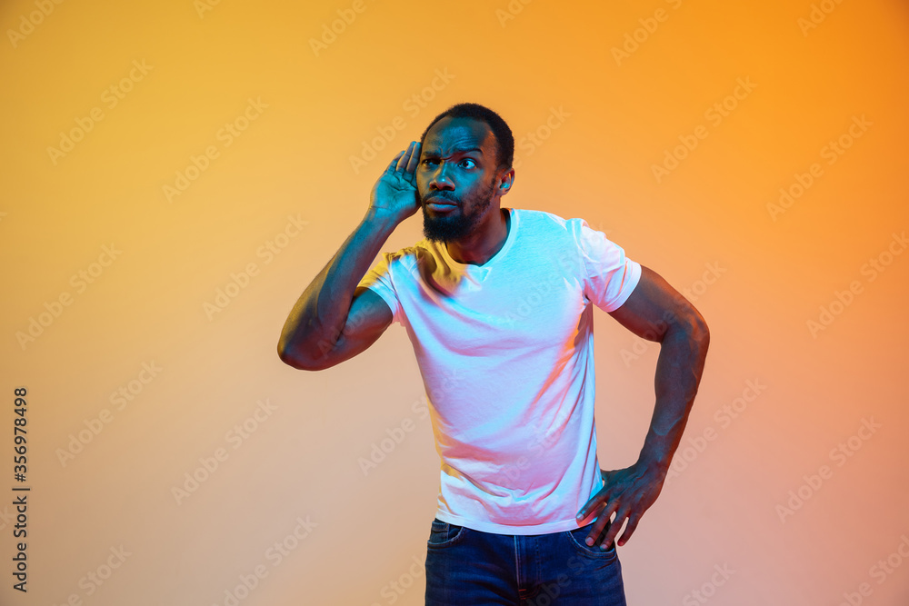 Listen to secrets with hand on ear. African-american man's modern portrait on gradient orange studio background in neon. Beautiful afro model. Concept of human emotions, facial expression, sales, ad.