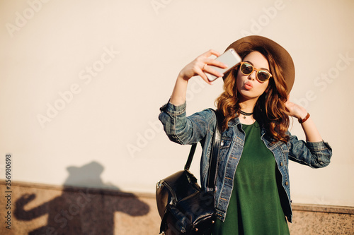 Brunette girl in sunglasses and hat making duck face, sending kiss at camera and taking self portrait at smart phone.Beautiful and funny woman posing, walking at street. Concept of street fashion. photo