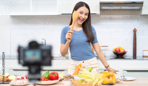 Social media concept. Happy woman standing in kitchen  using camera and recording online video. Happy asian woman vlogger broadcasting live video online teaching cooking food in kitchen at home