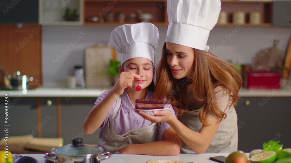 Mother and daughter in hats decorating cake with cherry on domestic kitchen Mother and daughter in hats decorating cake with cherry on domestic kitchen