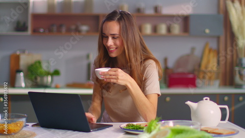 Woman having video chat on laptop at kitchen. Girl drinking coffee at home