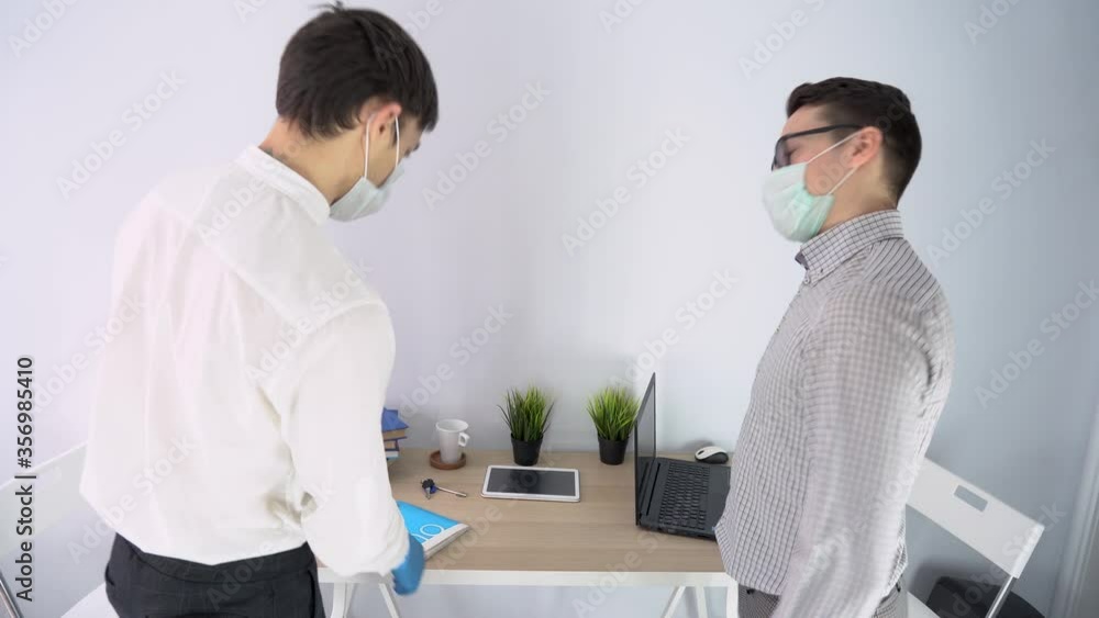 Two worker men wear shirts, protective medical face masks, meet in office room, greet, sit at table with laptop, plants, notebook. People interviewing for new job, talking, keep social distance indoor Two worker men wear shirts, protective medical face masks, meet in office room, greet, sit at table with laptop, plants, notebook. People interviewing for new job, talking, keep social distance indoor