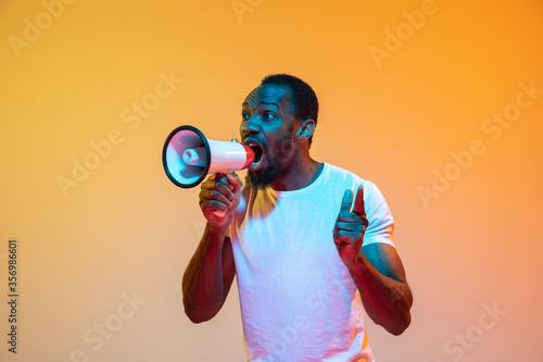 Shouting with megaphone. African-american man's modern portrait on gradient orange studio background in neon. Beautiful afro model. Concept of human emotions, facial expression, sales, ad. Copyspace. © master1305
