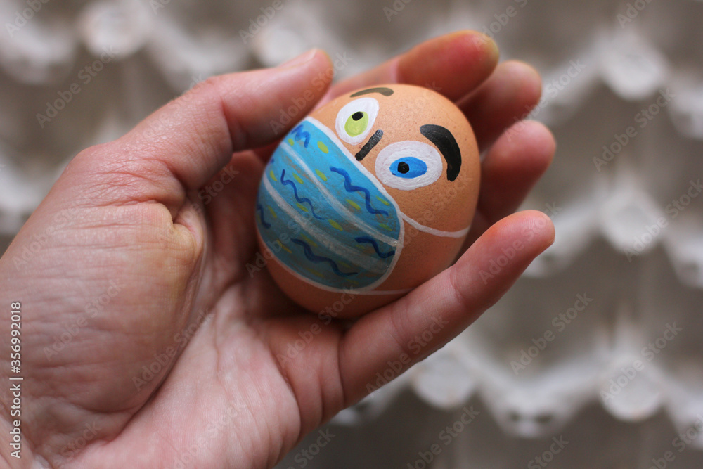 Painted Easter egg like human face with protective masks in the human hand, eggs carton on the background Painted Easter egg like human face with protective masks in the human hand, eggs carton on the background