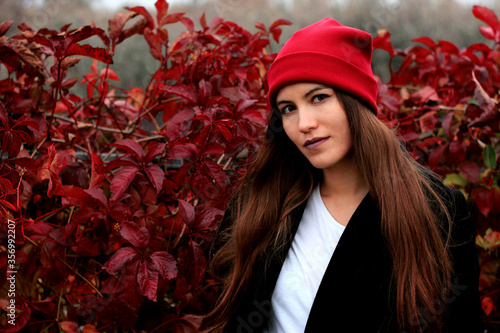 The girl with long hair on the background of the fence overgrown with ivy. A beautiful girl in a red hat, in a black coat against a red hedge.