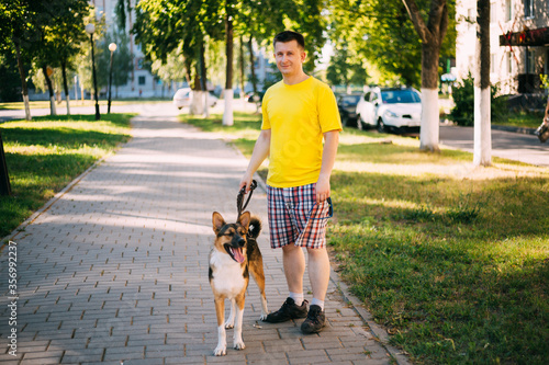 Young Caucasian Man Standing With Funny Mixed Breed In Park Walkway. Dog Walking On Leash