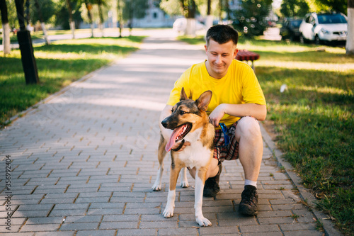 Young Caucasian Man Sitting With Funny Mixed Breed In Park Walkway. Dog Walking On Leash. Man Wearing A Yellow Jersey And Light Shorts