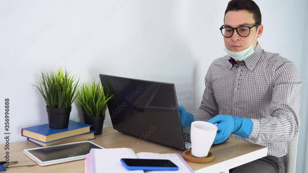 Young nice Caucasian man wear gray shirt, medical blue rubber gloves, black glasses, remote works, use laptop at home. Male want to drink, lowers face mask, takes cup on table, continues. Workplace. Young nice Caucasian man wear gray shirt, medical blue rubber gloves, black glasses, remote works, use laptop at home. Male want to drink, lowers face mask, takes cup on table, continues. Workplace.