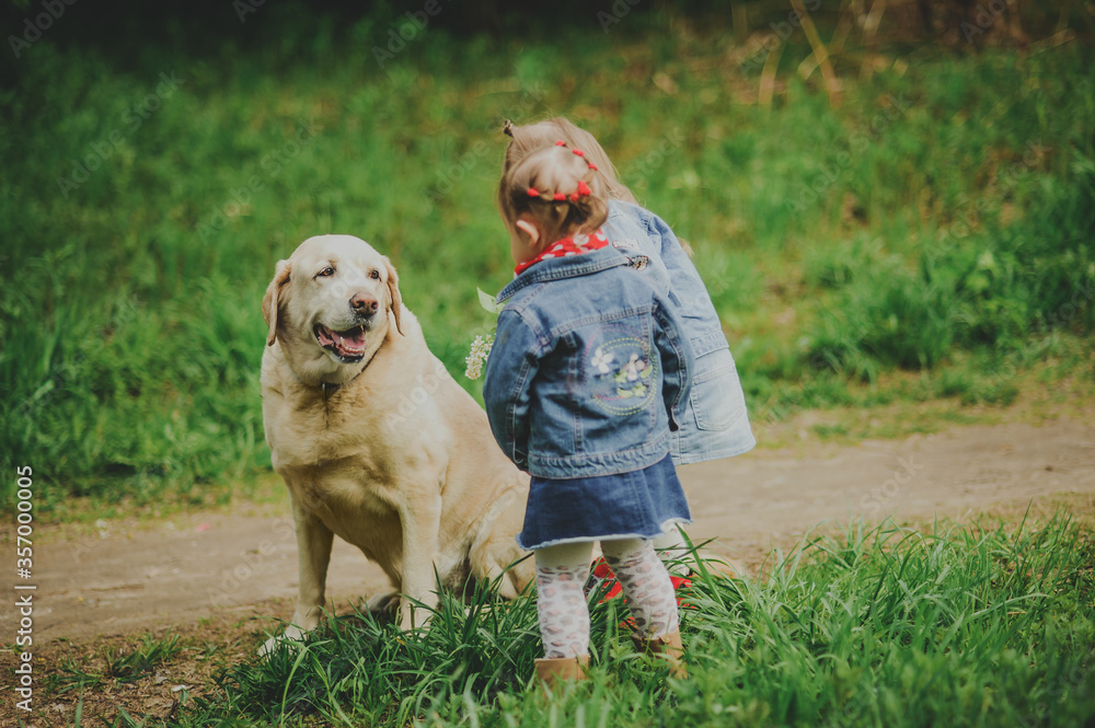 Two little girls in denim clothes pet a Labrador dog.