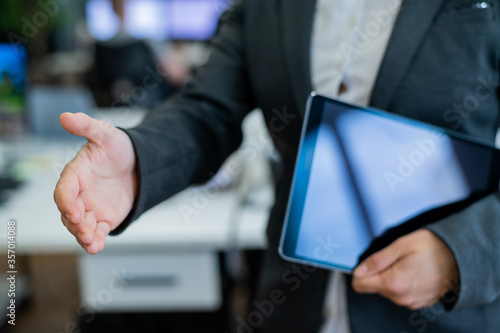 Business woman in a jacket and white shirt holding out a hand for a greeting. A faceless business woman holds a digital tablet and shakes her hand as an agreement on a successful transaction. © Михаил Решетников