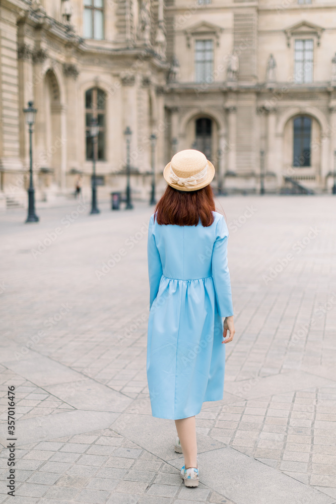 Back view of beautiful elegant young lady in hat and blue dress, walking near the vintage building in European city