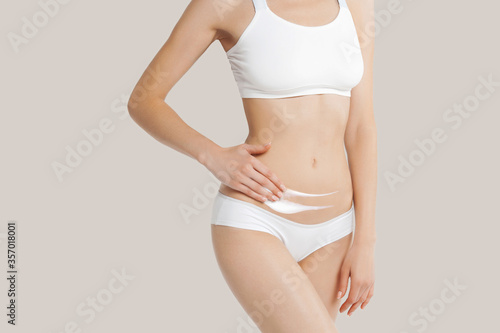 Slim young woman in underwear apply cream to belly on beige background