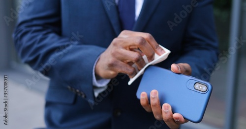 Close up of smartphone in hands of African American businessman in suit and tie. Man cleaning phone with disinfecting napkin. Cellphone disinfection outdoor.