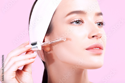 Young beautiful woman with clean perfect skin uses age-sensitive serum in pipette on pink background