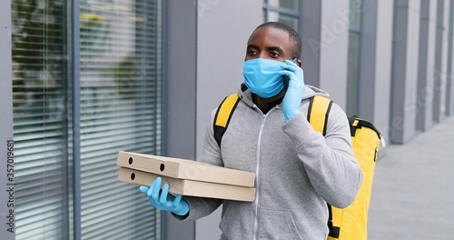 African American young deliveryman in medical mask and gloves walking at street, talking on mobile phone and looking for address. Male courier with boxes having a walk and speaking on cellphone.