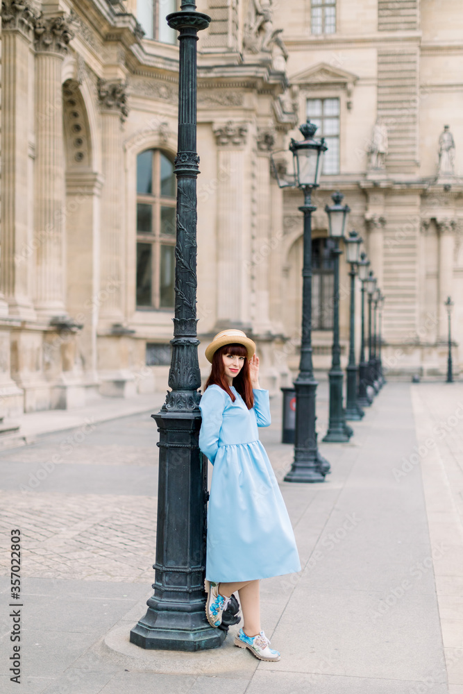 Full length portrait of beautiful young smiling Caucasian woman in elegant blue dress and hat, standing near the old vintage building and leaning on vintage street lamp