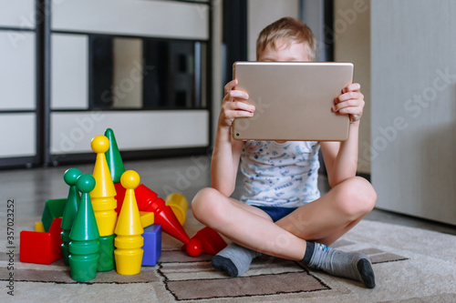 The boy is in the game room, next to toys, but does not play, but looks at the tablet