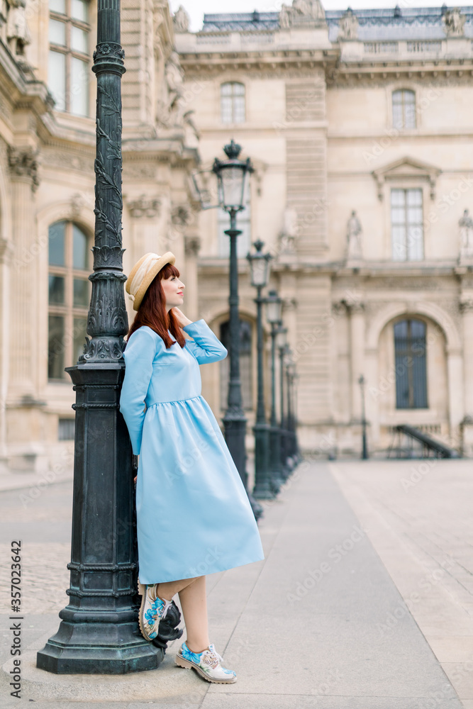 Fototapeta premium Side view of beautiful young woman in elegant blue dress and hat, standing near the vintage building outdoor and leaning on vintage street lamp and looking up